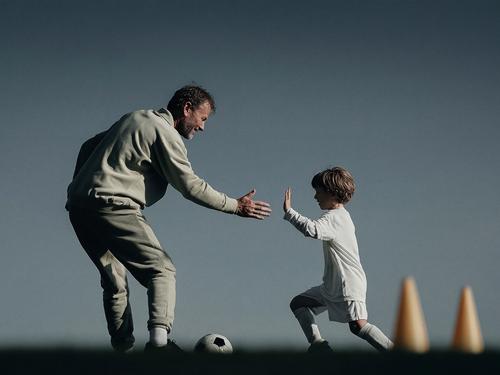 주황색 콘이 있는 축구장에서 흰색 운동복을 입은 어린 소년과 하이파이브를 하며 미소 짓는 나이든 코치 또는 멘토