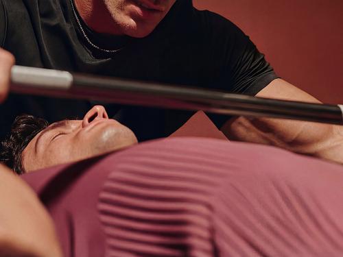 Close-up de um auxiliar ou coach ajudando uma pessoa deitada no banco de supino, levantando uma barra.