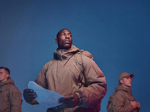 파란색 배경에 다른 사람들과 함께 일렬로 서서 군용 또는 전술 장비와 배낭을 착용하고 지도를 든 채 집중하고 있는 남성