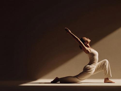 측면에서 드라마틱한 조명이 비치는 요가 스튜디오에서 밝은 색상의 요가복을 입은 채 초승달 런지 자세(안자네야사나)를 취하고 있는 여성